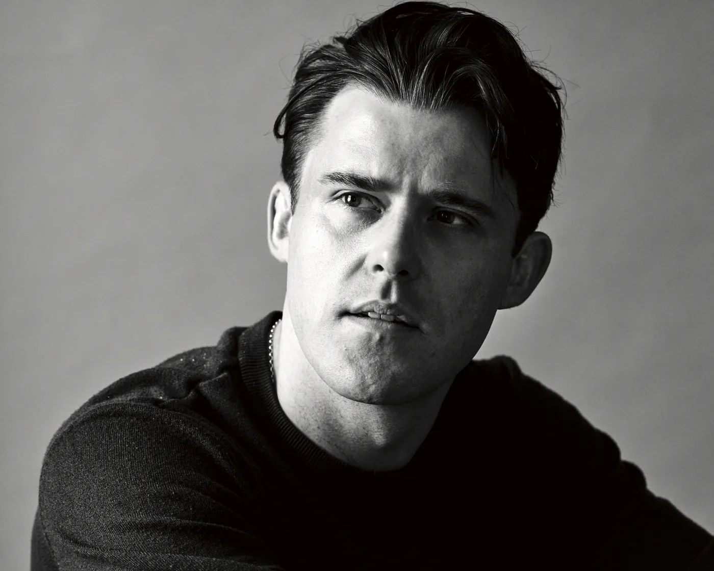 A black and white studio portrait of a man in a dark jumper against a plain background