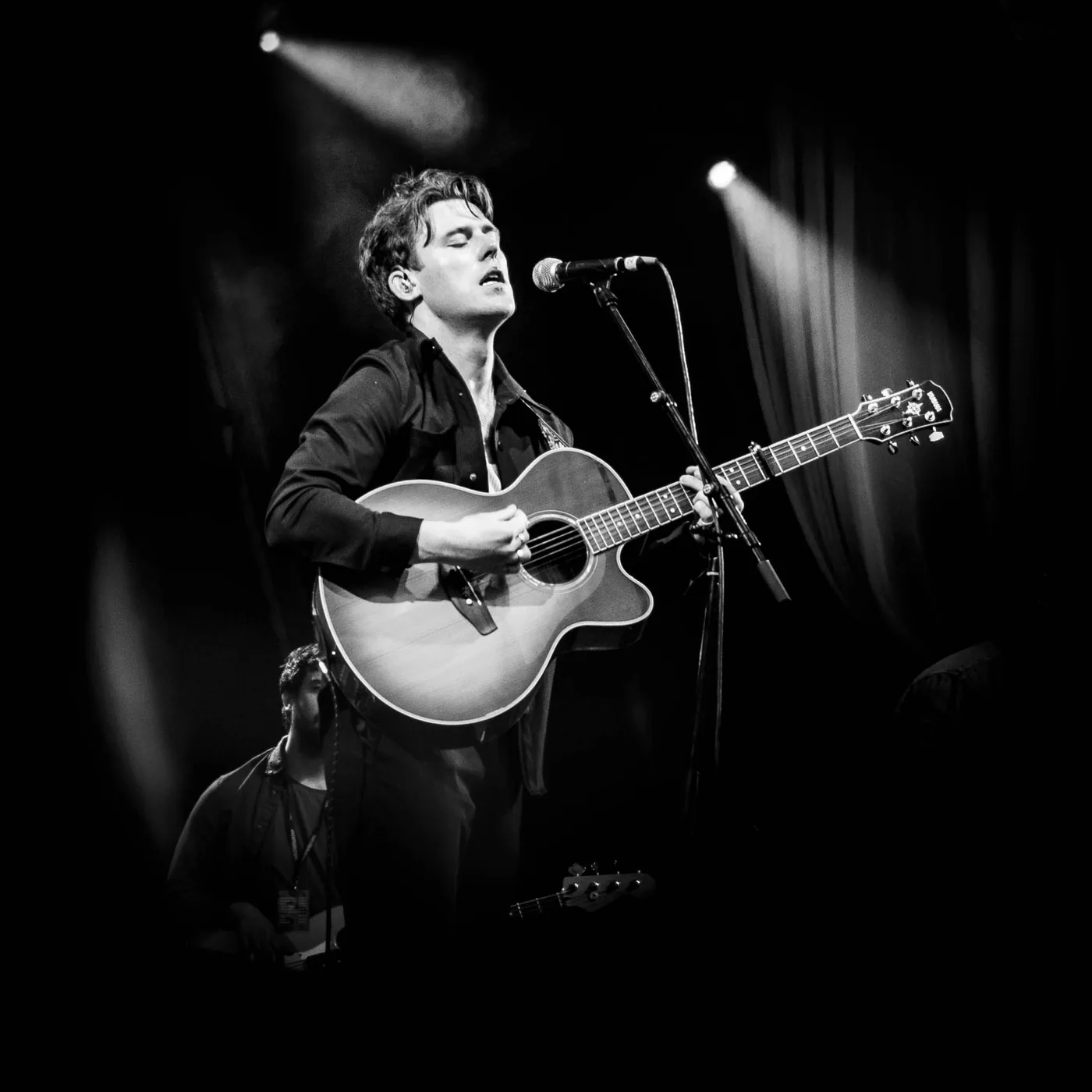 A black and white photograph of a man playing an acoustic guitar at a microphone under a single spotlight