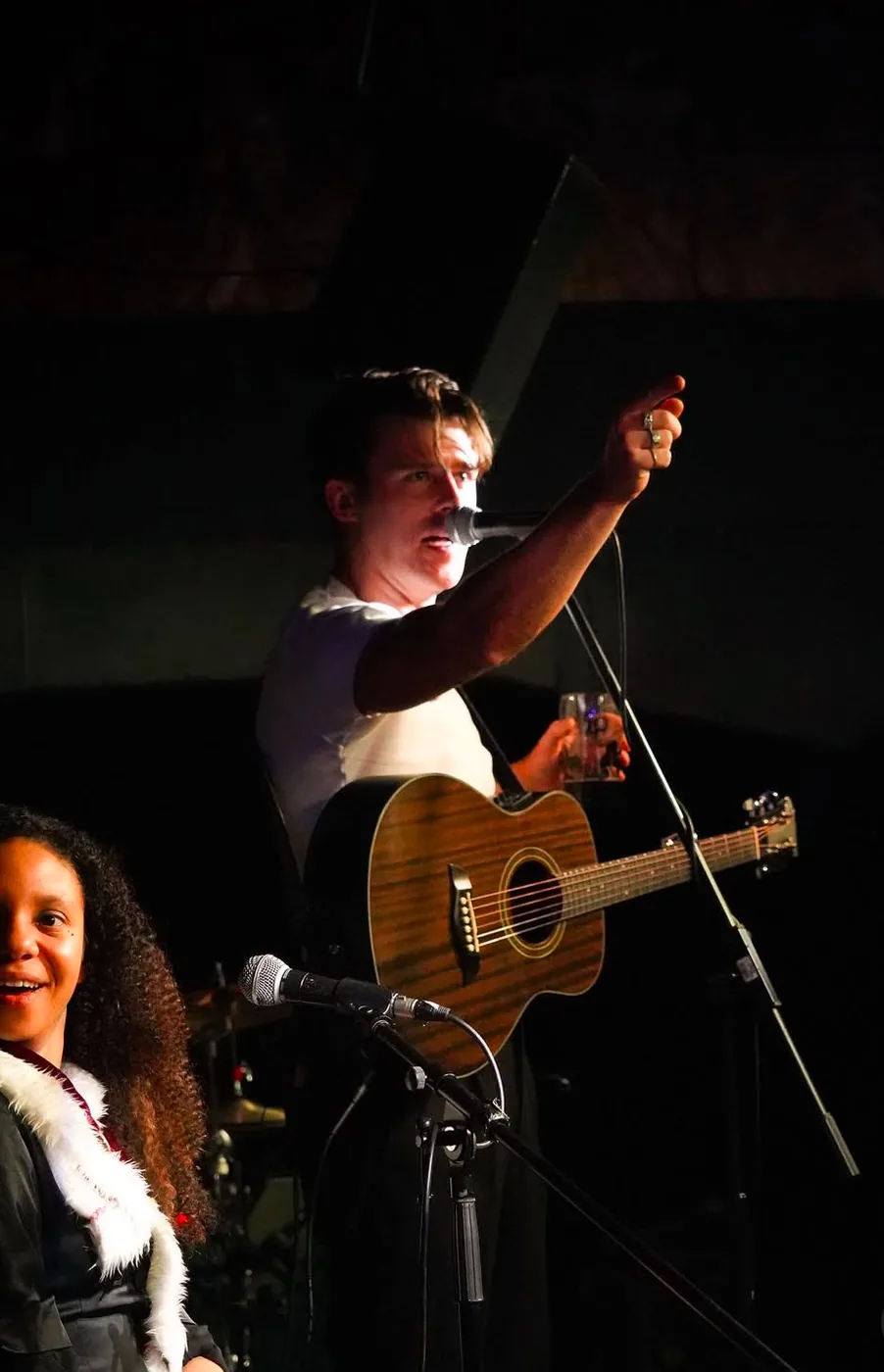 A singer with an acoustic guitar reaching an arm above his head at the microphone, an audience member in front of the stage
