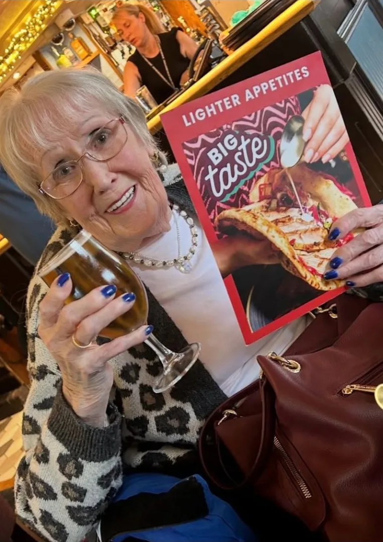 A smiling woman raises a glass of beer and holds up a pub menu at a Cabaret Lunch Club